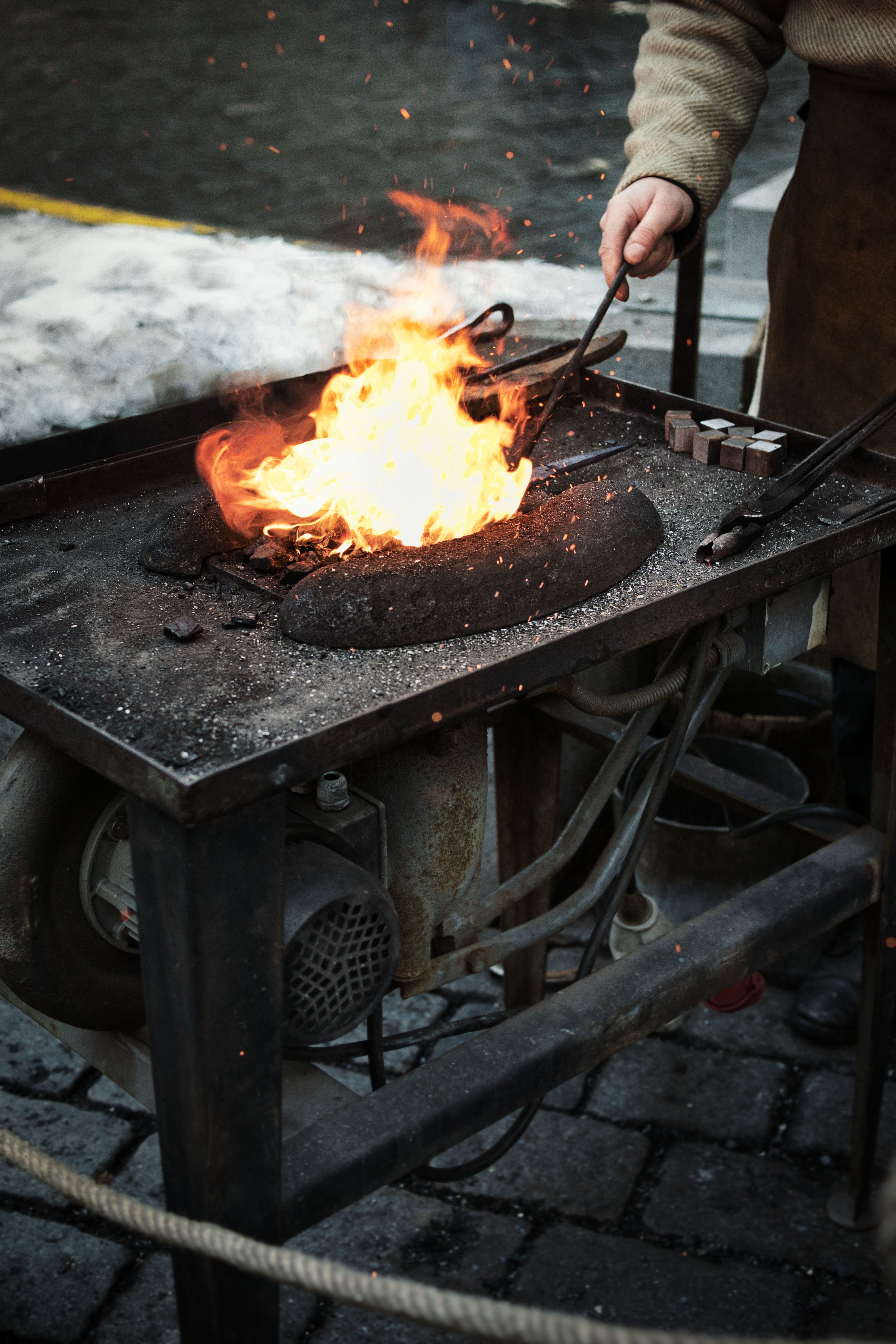 A blacksmith tending to a blazing forge outdoors, showcasing craftsmanship and traditional metalworking.