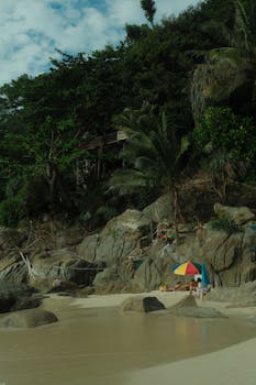 A tropical beach scene with rocks, greenery, and a colorful umbrella under clear skies.