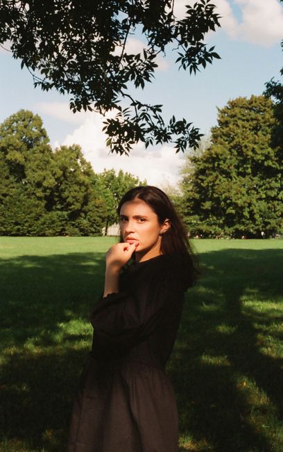 A young woman poses in a sunlit park, surrounded by lush greenery.