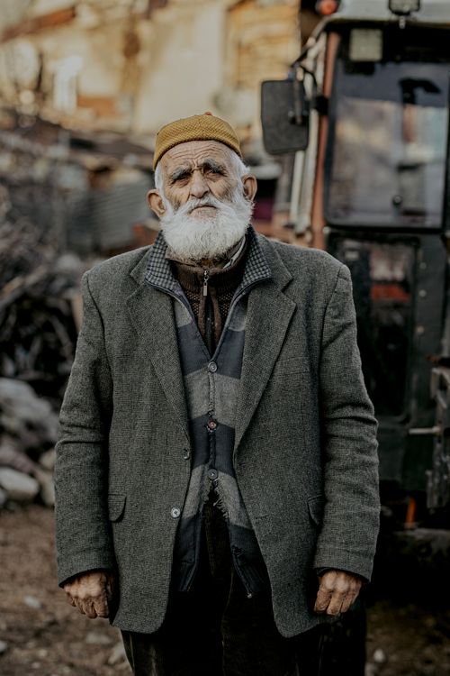 Free Portrait of a senior man with a beard, standing outdoors next to a rustic tractor in a rural setting. Stock Photo