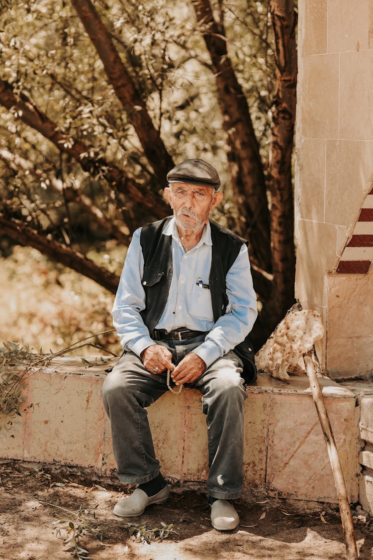 Elderly Man In Black Vest And Shirt Sitting On Concrete