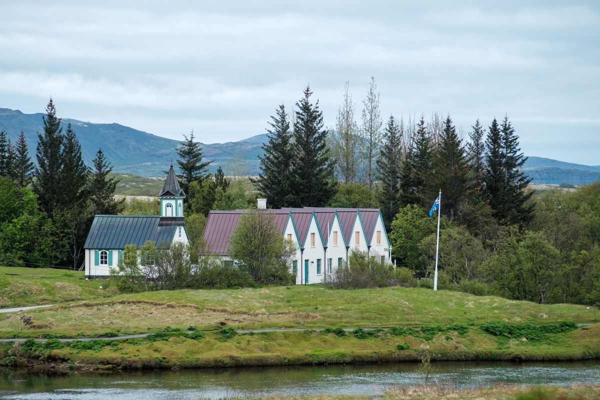 Small white church with a dark spire beside a turf-and-timber farmhouse at Þingvellir, trees and a flagpole nearby, a river in the foreground.
