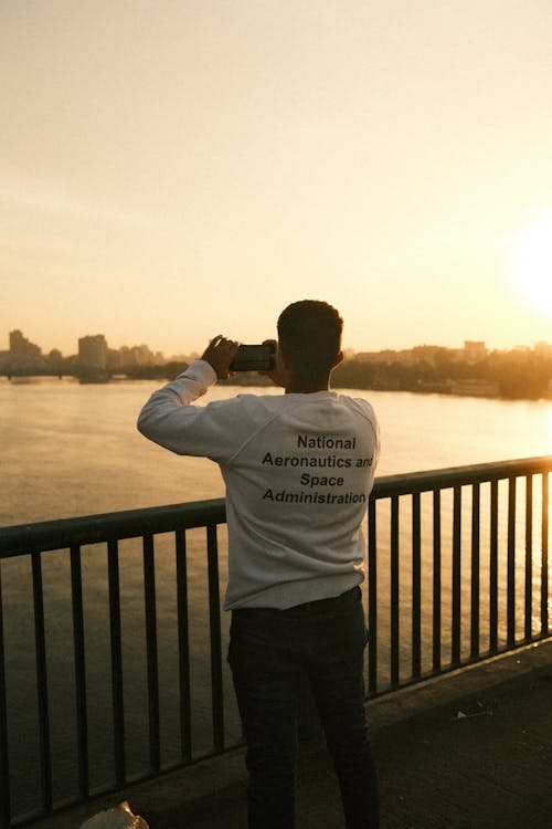 Man Standing by Railing over River and Taking Pictures at Sunset