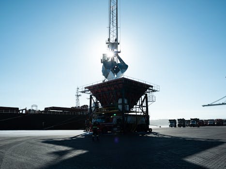 An industrial port scene showing a large crane with sunlight behind, surrounded by trucks.
