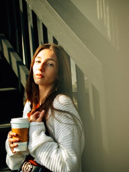 A young woman in a white sweater enjoys a moment with coffee in a sunlit urban environment.
