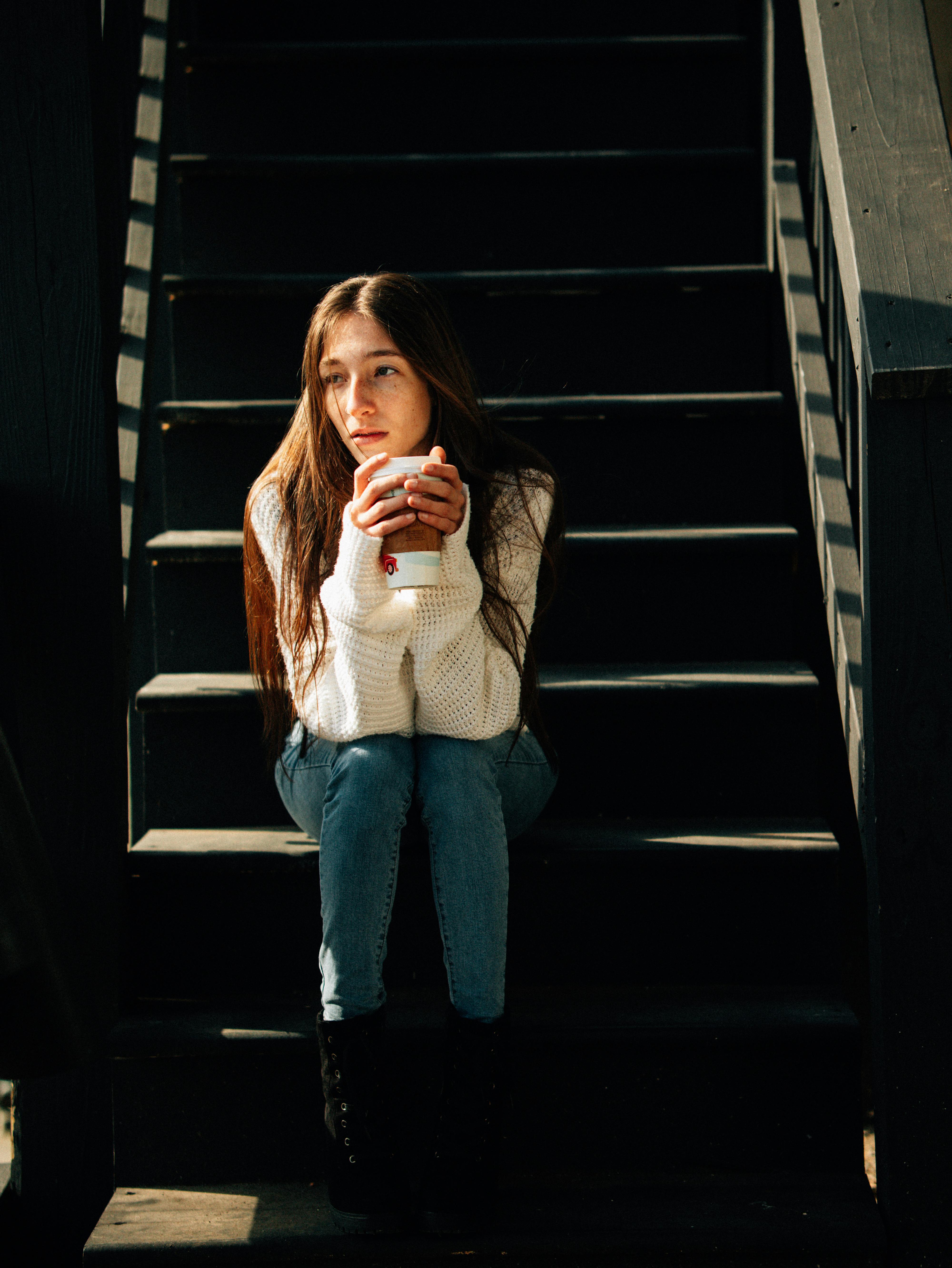 A young woman in a white sweater holds a cup, sitting on shadowed stairs, deep in thought.