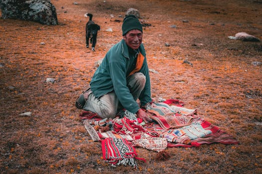 Man in rural Peru displays handwoven Andean textiles on grassland. A dog roams nearby.