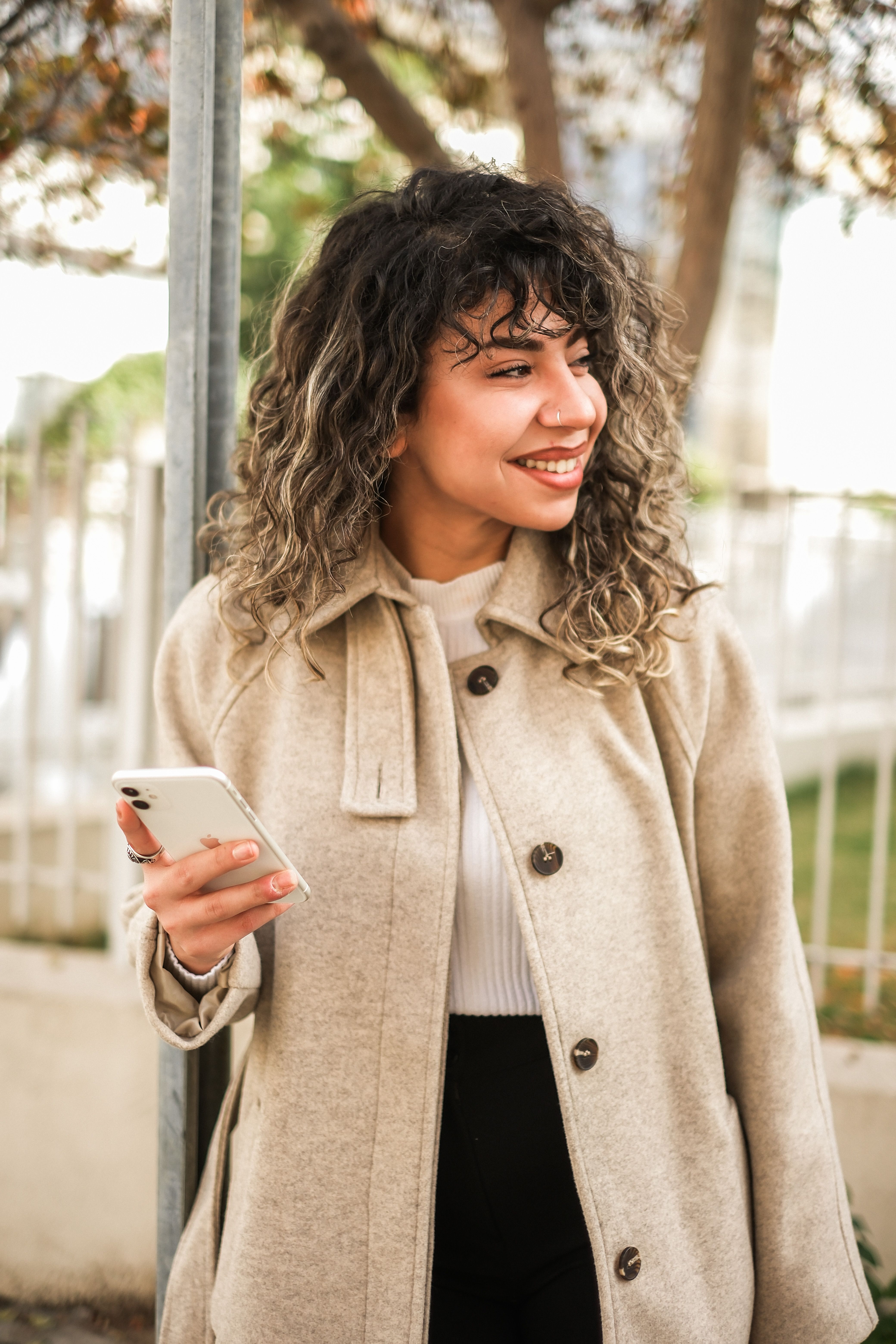 A fashionable woman with curly hair smiles while holding a smartphone outdoors.