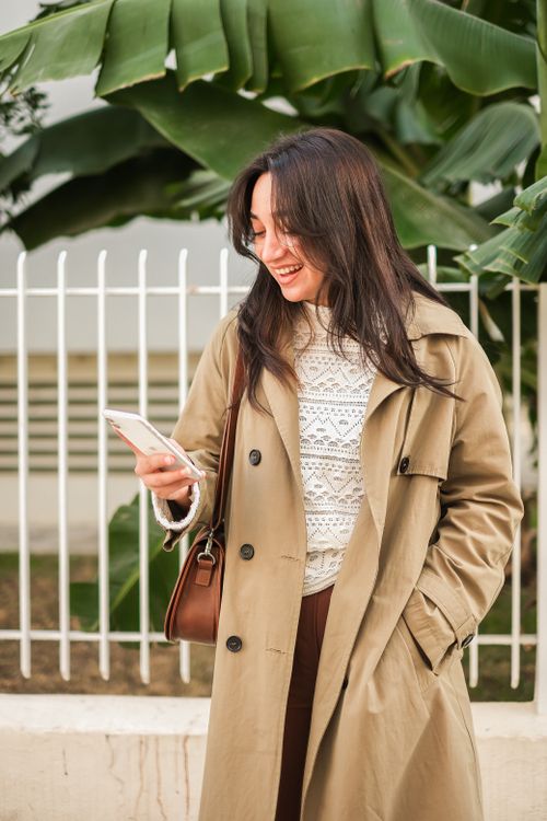Smiling woman in beige coat checking her phone outdoors