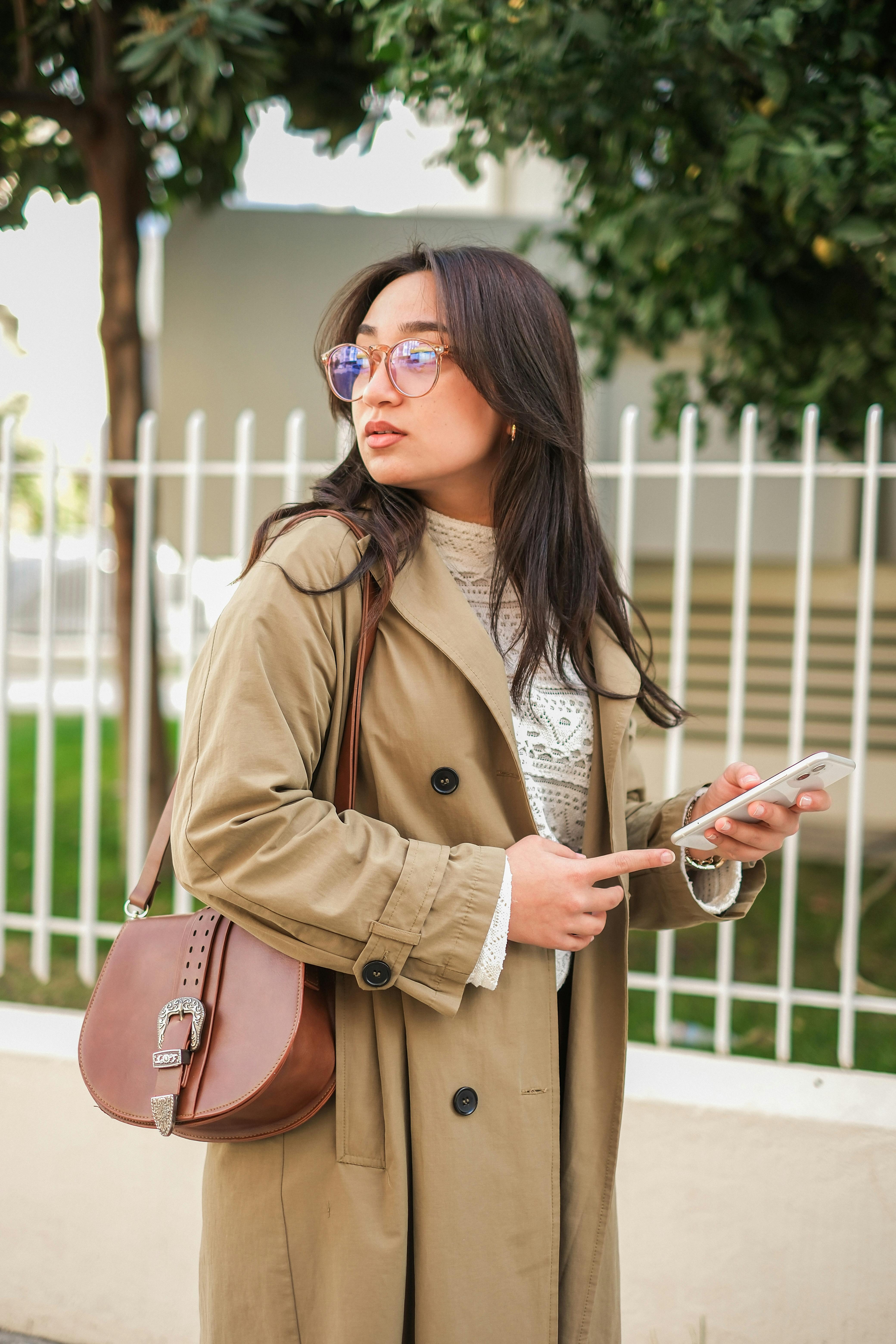 Fashionable woman outdoors with a trench coat and handbag, using her smartphone.