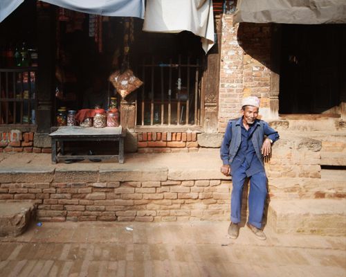 Free An elderly man in traditional clothing sits outside a rustic shop in sunlight. Stock Photo