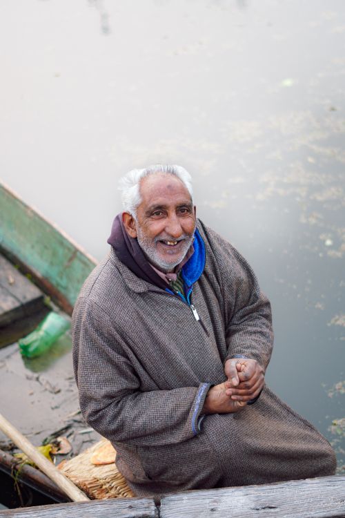 Free Elderly man with gray hair and beard stands smiling by a boat on serene water. Stock Photo