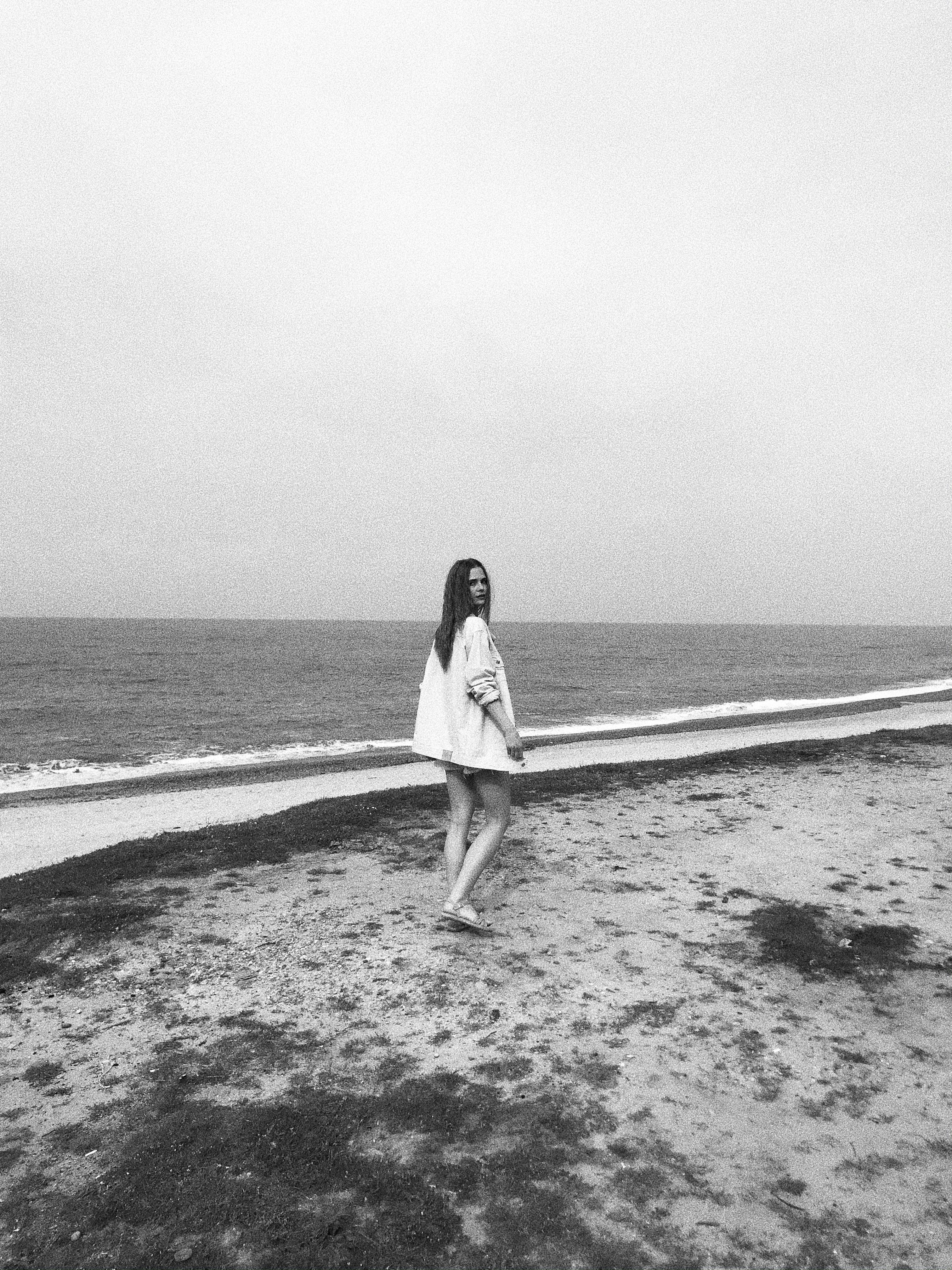 A woman walks leisurely on a quiet beach, gazing at the sea, captured in black and white.