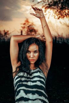 Portrait of a young woman striking a pose with arms raised against a dramatic sunset background.