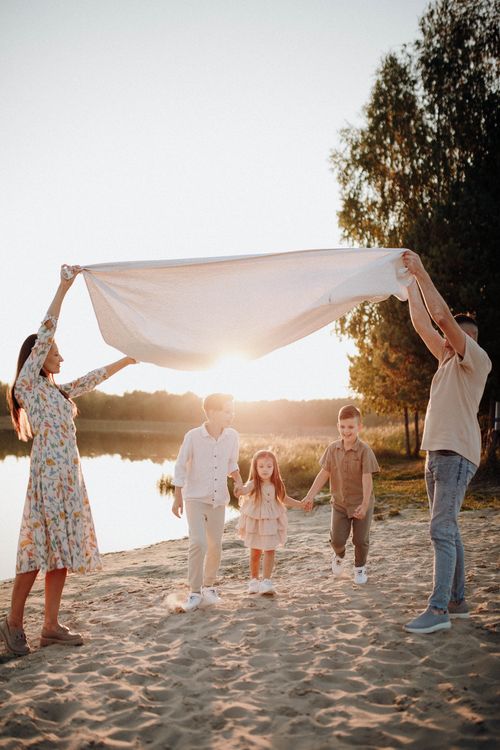 Free Family of five playing with a blanket at a lakeside beach during a sunny day, depicting joy and togetherness. Stock Photo