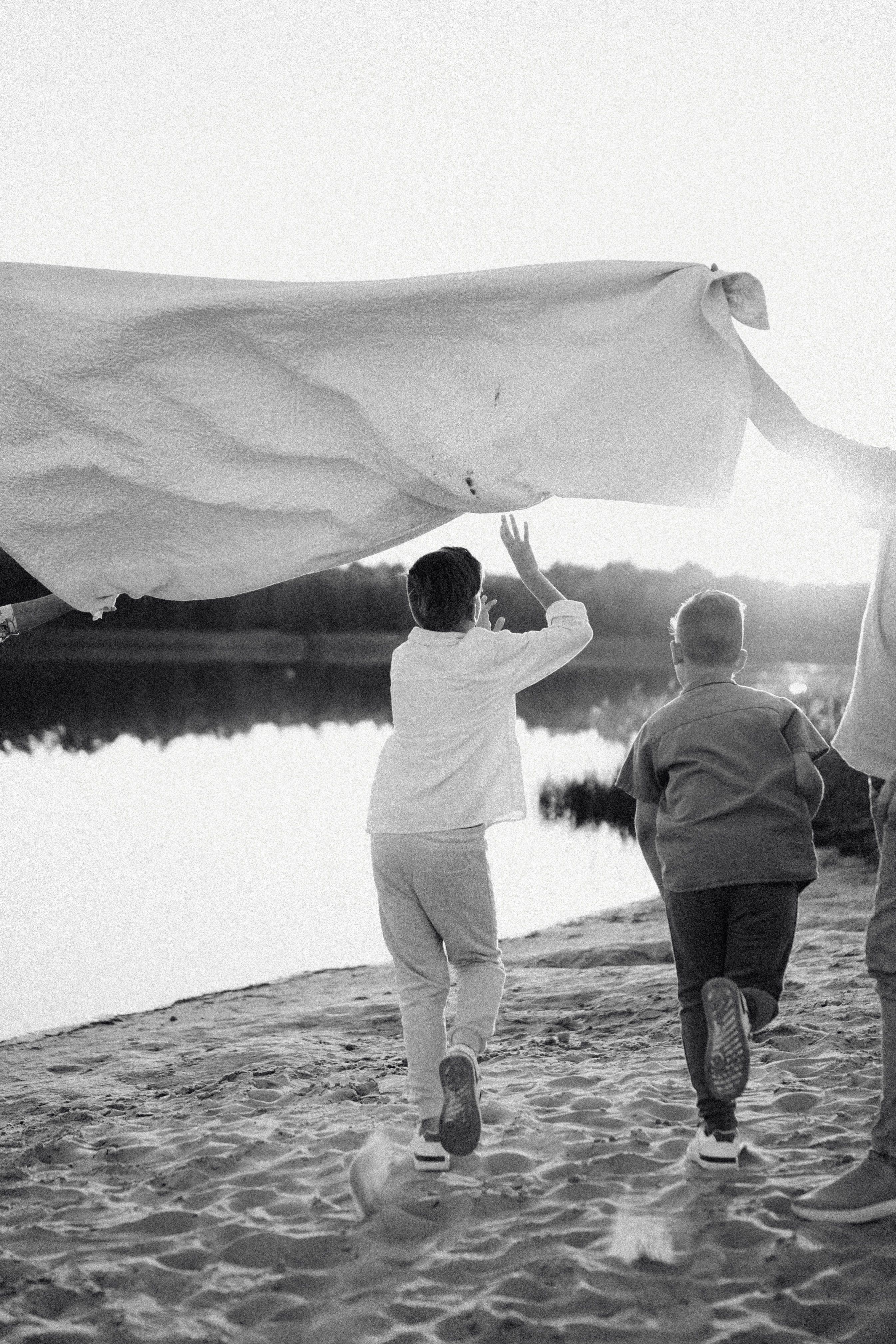 Boys Run on Sandy Beach beside Lake under Blanket · Free Stock Photo