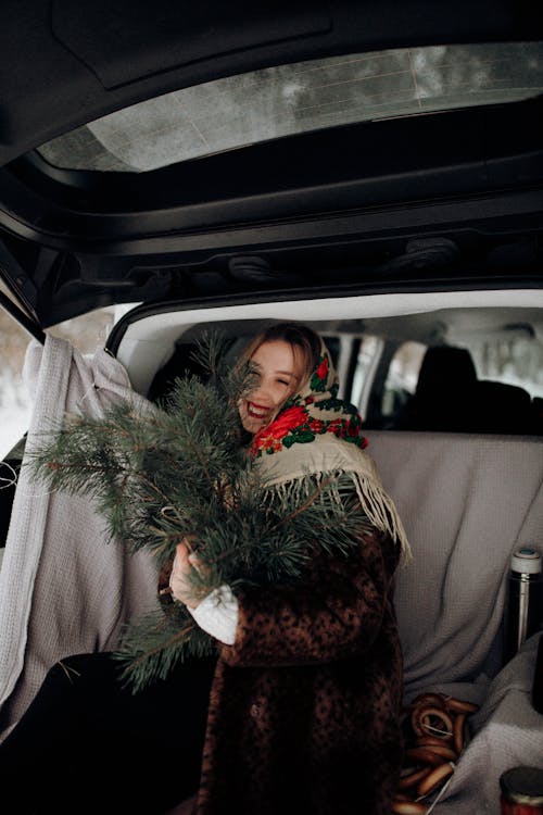Smiling Woman Sitting with Christmas Tree in Car Trunk