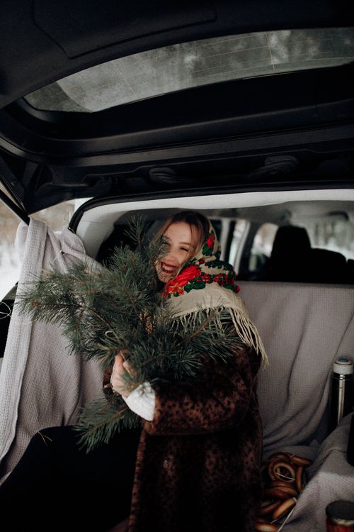 Free Smiling woman in cozy attire holds evergreen branches in car trunk, winter setting. Stock Photo