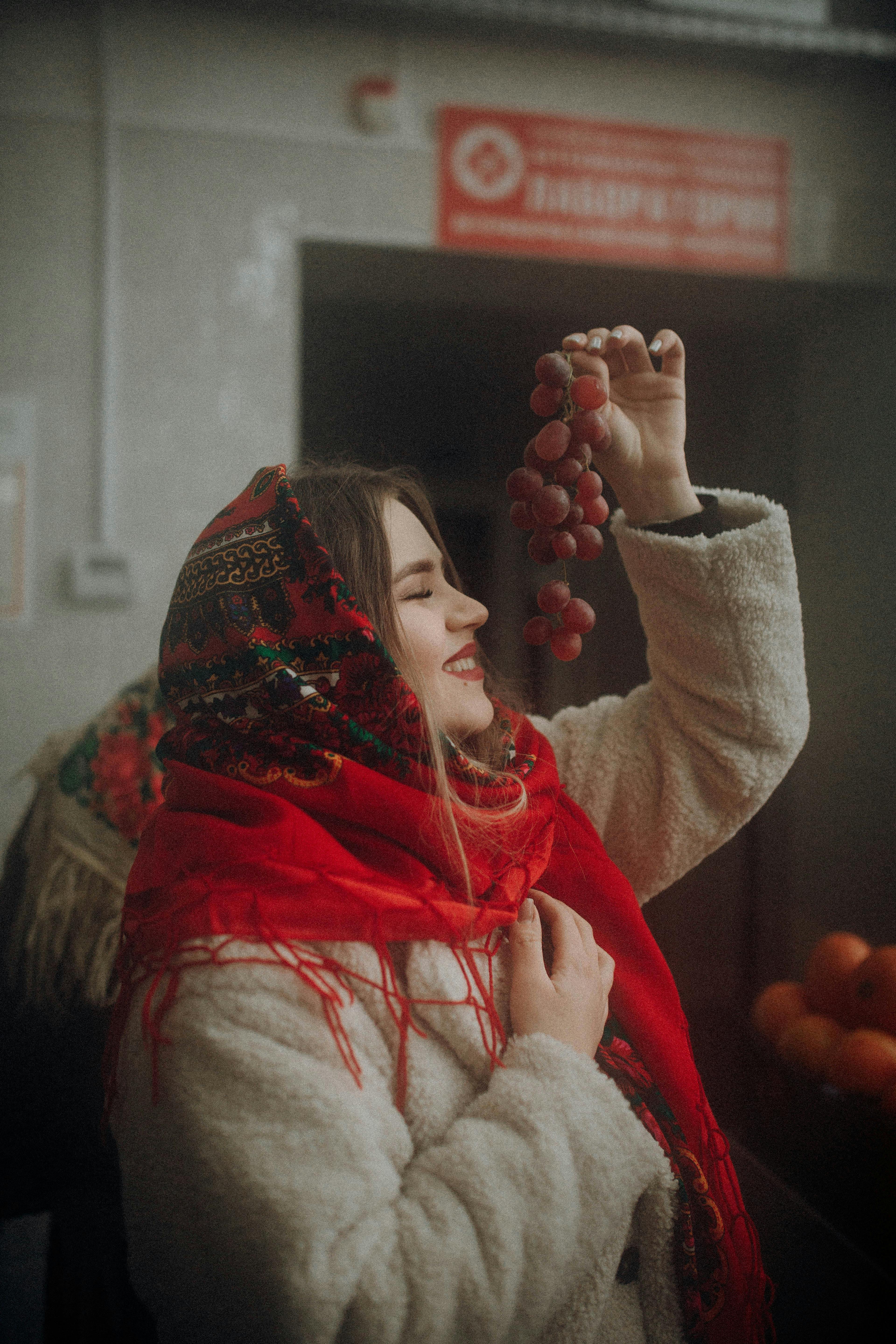 Smiling young woman with a red scarf holds grapes in festive winter attire indoors.