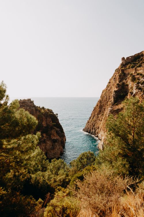 Free Vibrant view of rocky cliffs meeting the blue sea, framed by lush greenery under a clear summer sky. Stock Photo