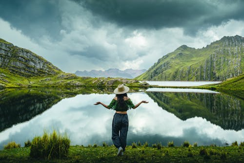 Woman in Hat Standing by Lake and Green Hills