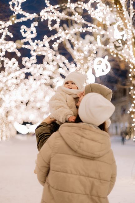 A mother lifts her bundled baby under captivating Christmas lights on a snowy winter night.