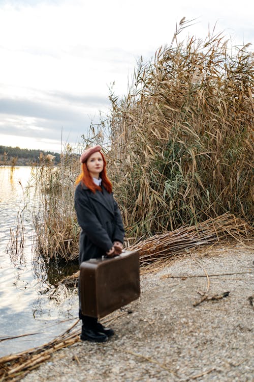 Redhead Woman in Coat Standing with Suitcase on Riverbank