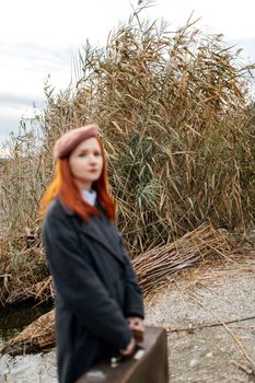 A stylish redhead woman in classic attire with a suitcase in a natural outdoor setting.