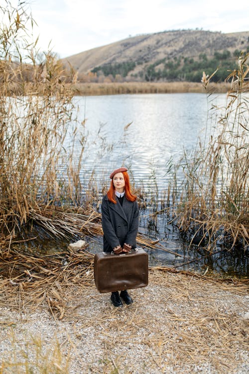 Redhead Woman Standing in Coat and with Suitcase on Riverbank