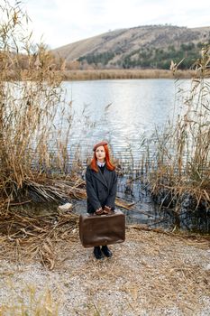 Red-haired woman in a beret by a river holding a vintage suitcase in autumn.