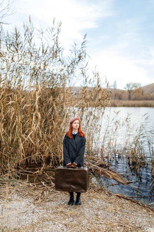 Young Red Haired Woman with Leather Suitcase Standing on Lakeside