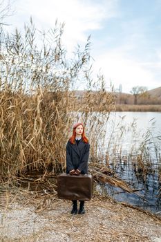 Red-haired woman holding a suitcase by a lake in autumn, blending fashion and nature.