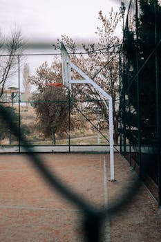 Outdoor basketball court with hoop surrounded by autumn foliage, viewed through a fence.