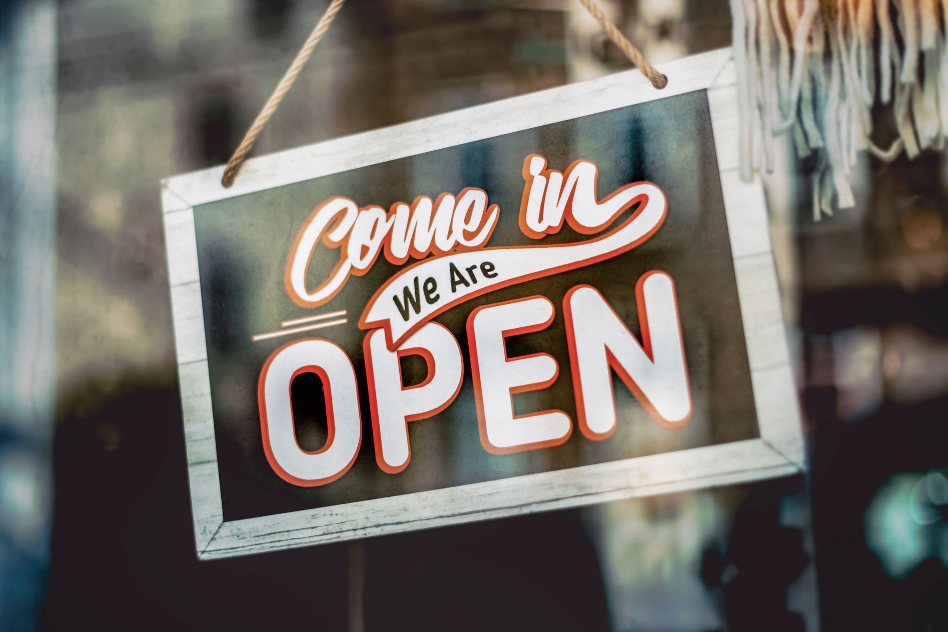 A welcoming 'Come in, We Are Open' sign hanging on a glass window of an urban shop.