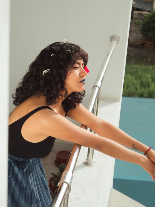 Free A woman with curly hair and floral hair decor leans on a balcony, enjoying the summer sunlight. Stock Photo