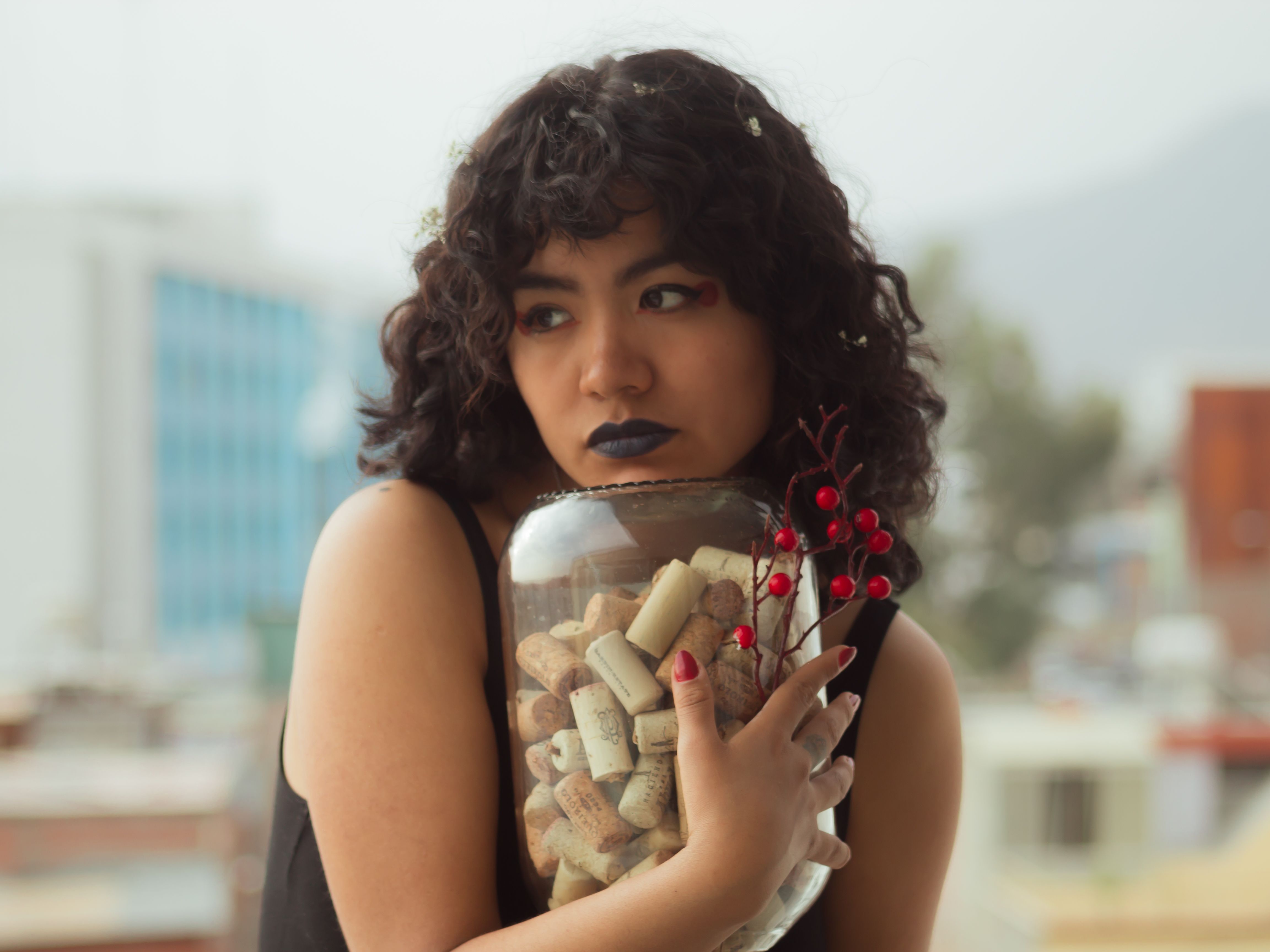 A woman with curly hair and blue lipstick holds a jar filled with corks and red berries.