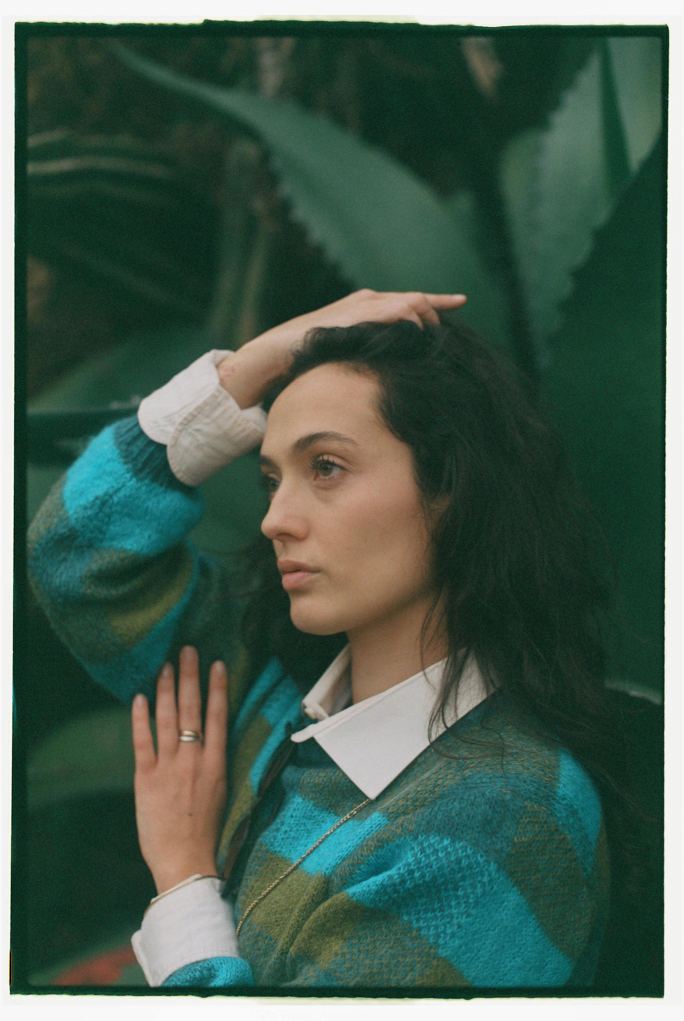 Portrait of a woman with long black hair in a vibrant sweater, set against lush greenery.