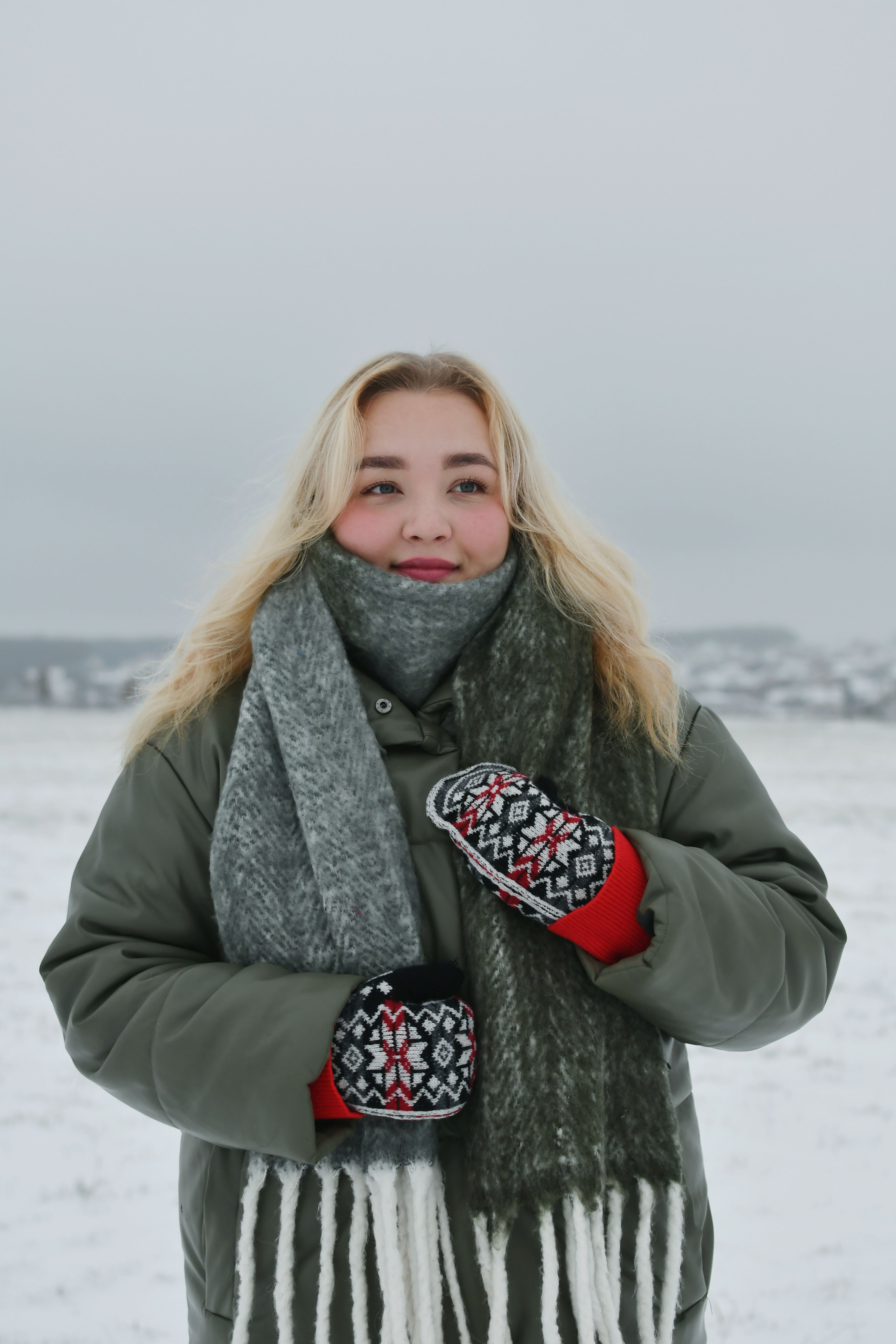 A young woman wearing winter attire stands in a snowy rural field, bundled in a scarf and mittens.