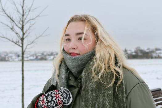 Blonde woman in warm coat and gloves enjoying a serene snowy landscape.