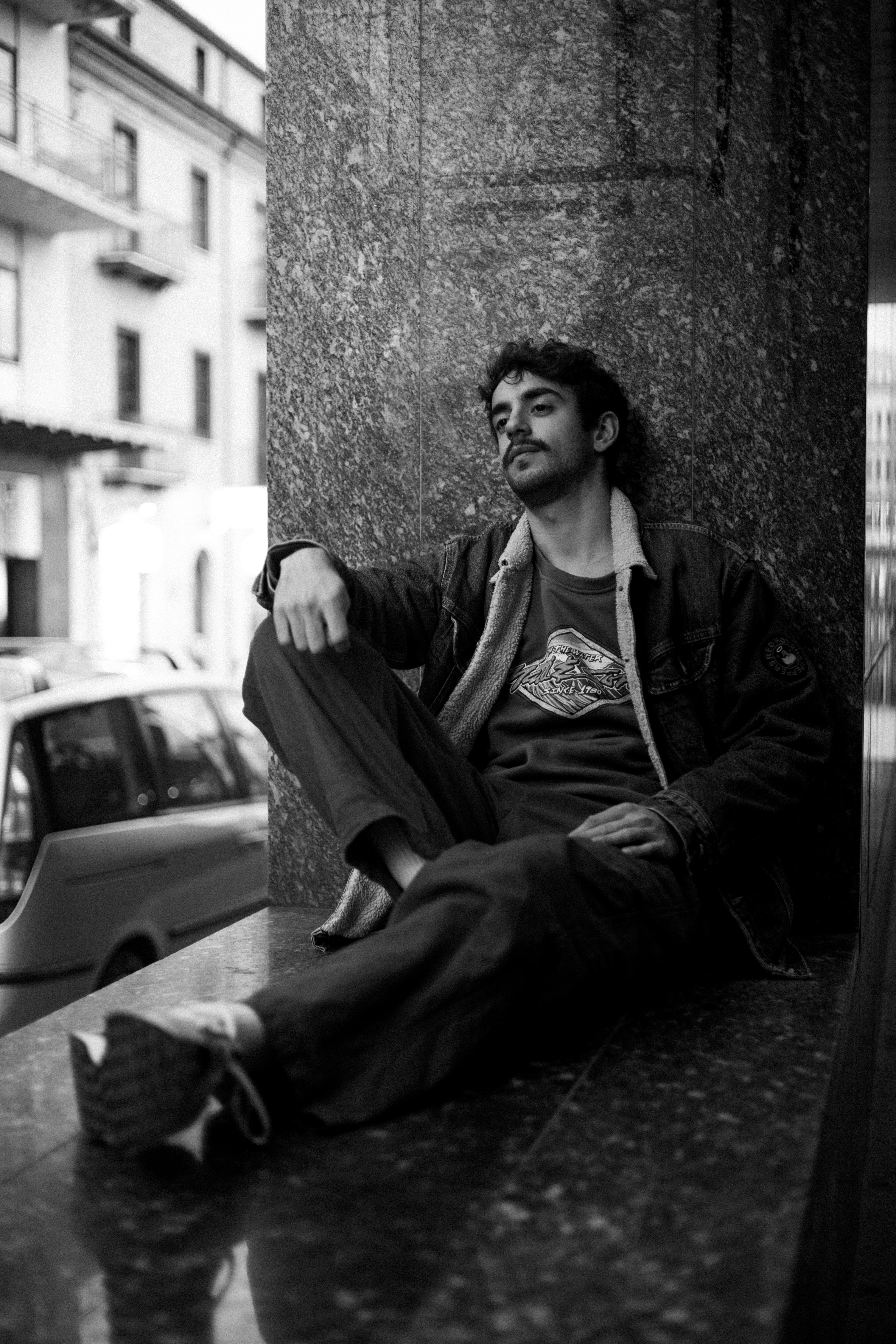 Stylish young man sits against a marble wall outdoors, exuding a relaxed vibe.