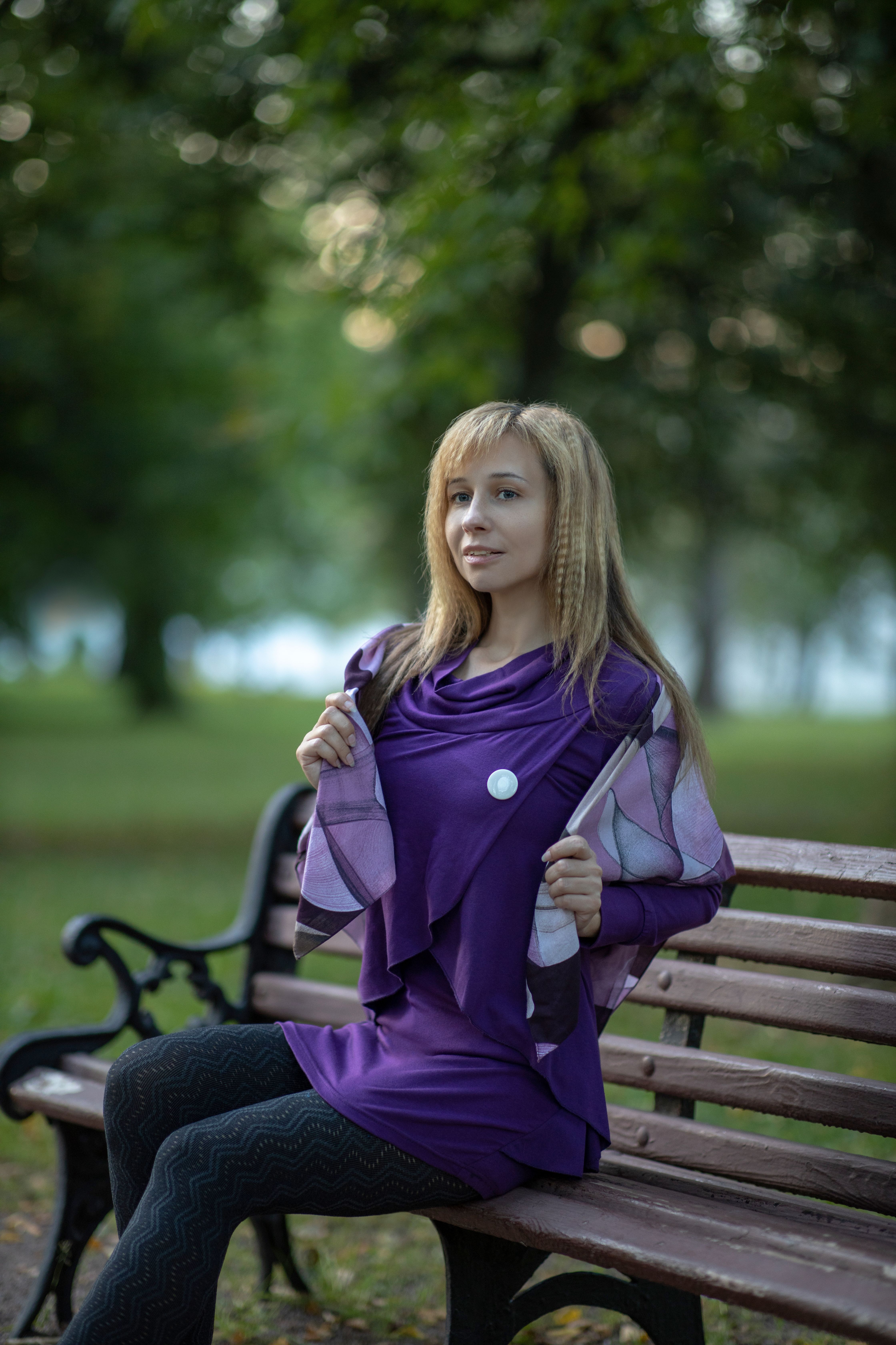 Woman in a purple dress poses on a bench in a lush green park setting.