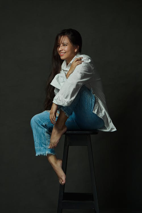 Woman in White Shirt and Jeans Sitting on Stool in Studio