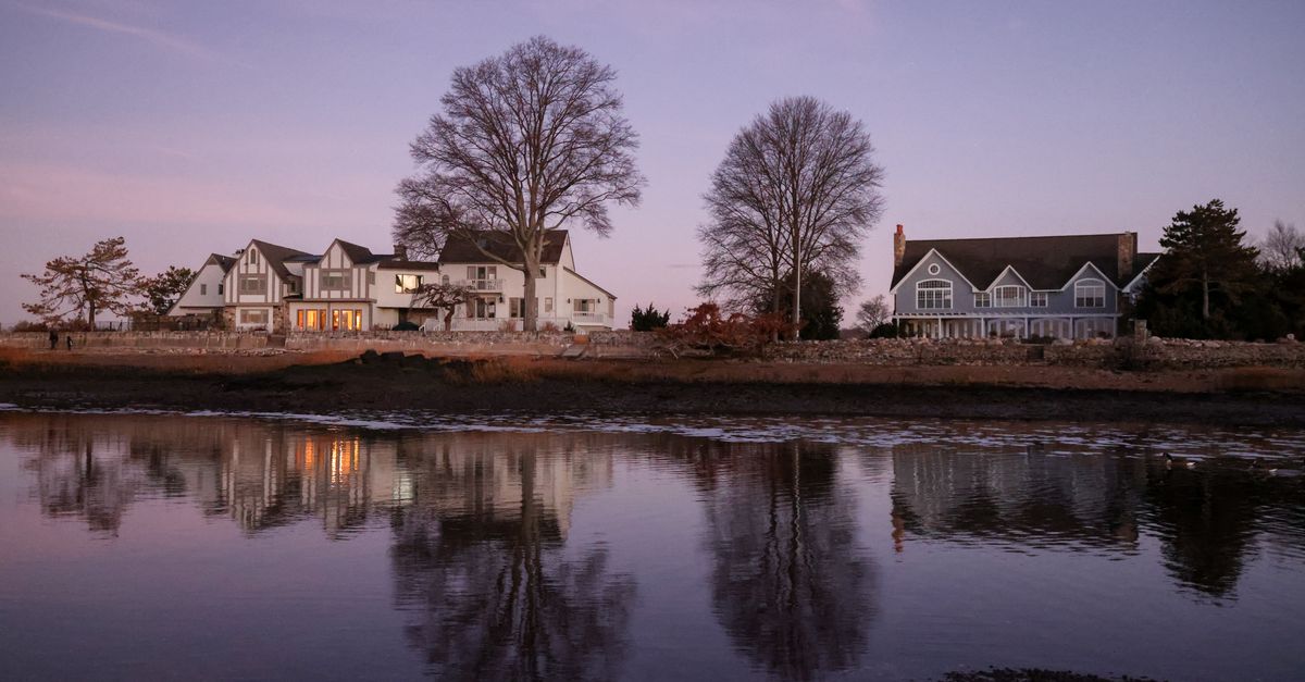 Charming waterfront homes reflect in the tranquil river at dusk, creating a serene rural scene.