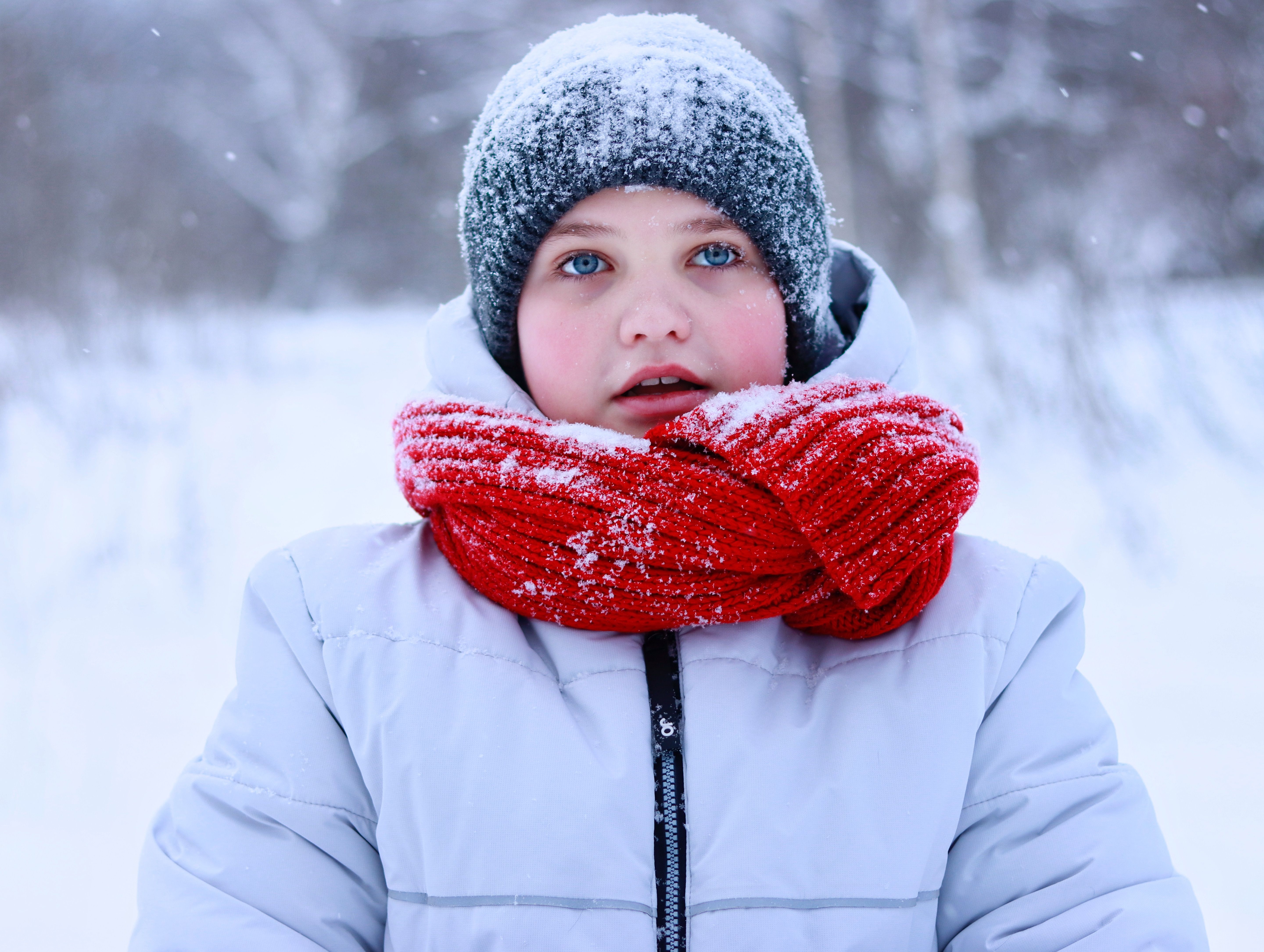 Jovem menina em cenário de inverno, vestida com roupas quentinhas e cobrindo-se do frio com cachecol e capuz, observando o ambiente nevado ao seu redor.