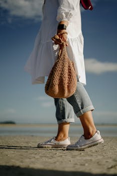 Woman walking on a sunny beach holding a knitted bag, wearing casual attire.