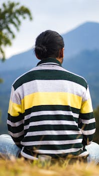 Back view of a man sitting outdoors wearing a striped sweater, enjoying mountain scenery.
