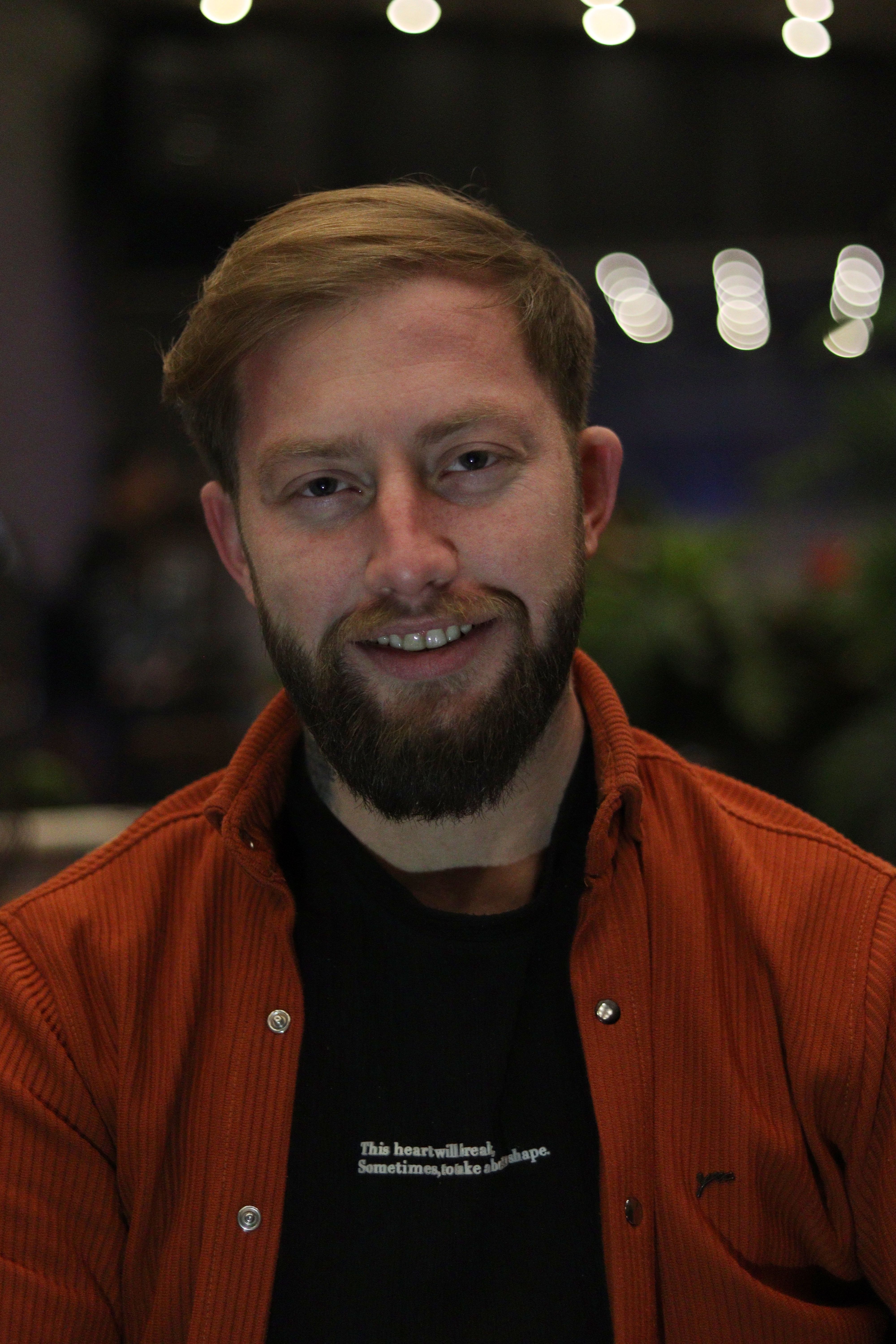A smiling man with a beard and short hair in an orange jacket indoors at night.