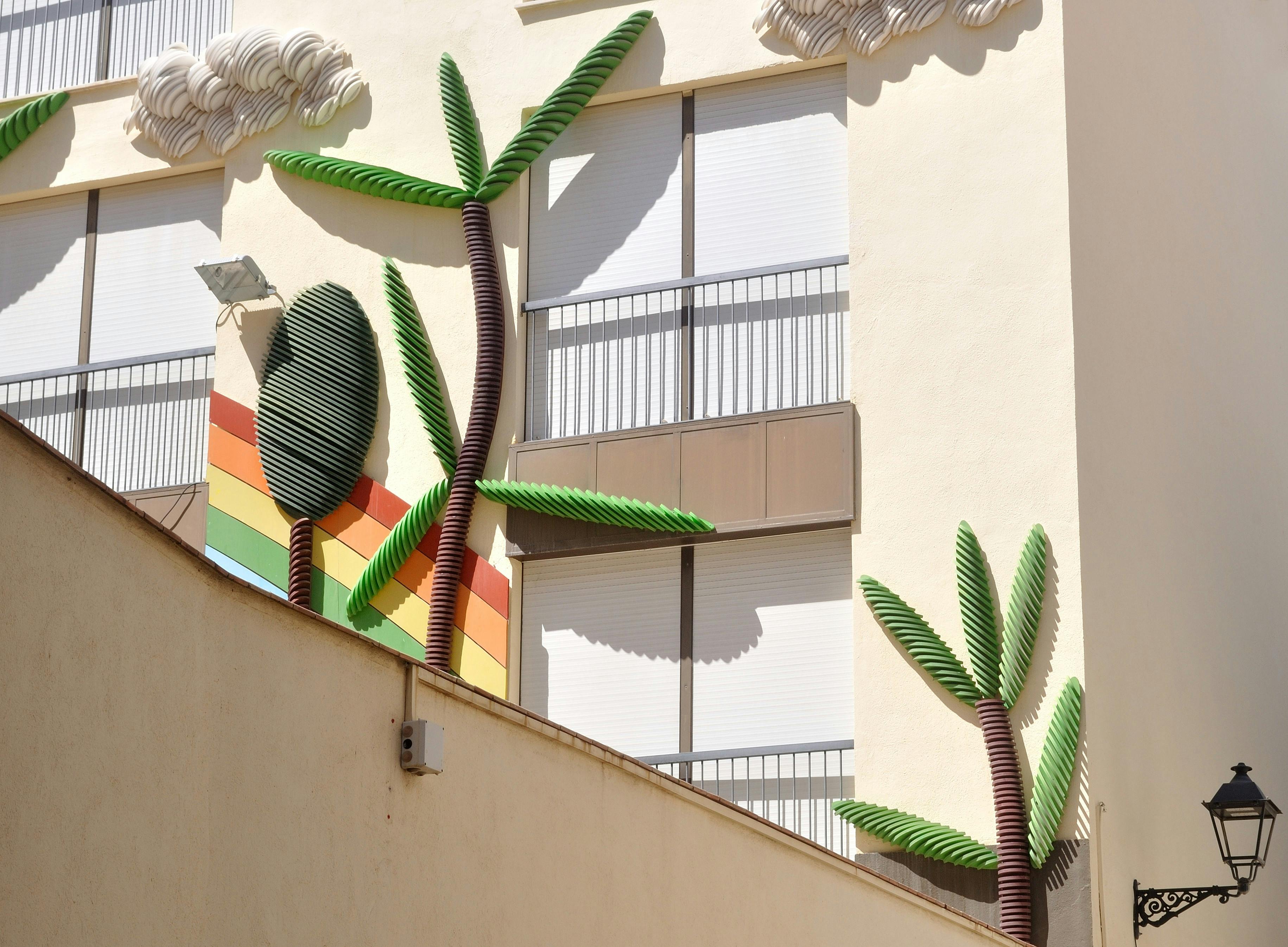 Vibrant urban wall art featuring 3D palm tree sculptures and rainbow patterns in a residential area.
