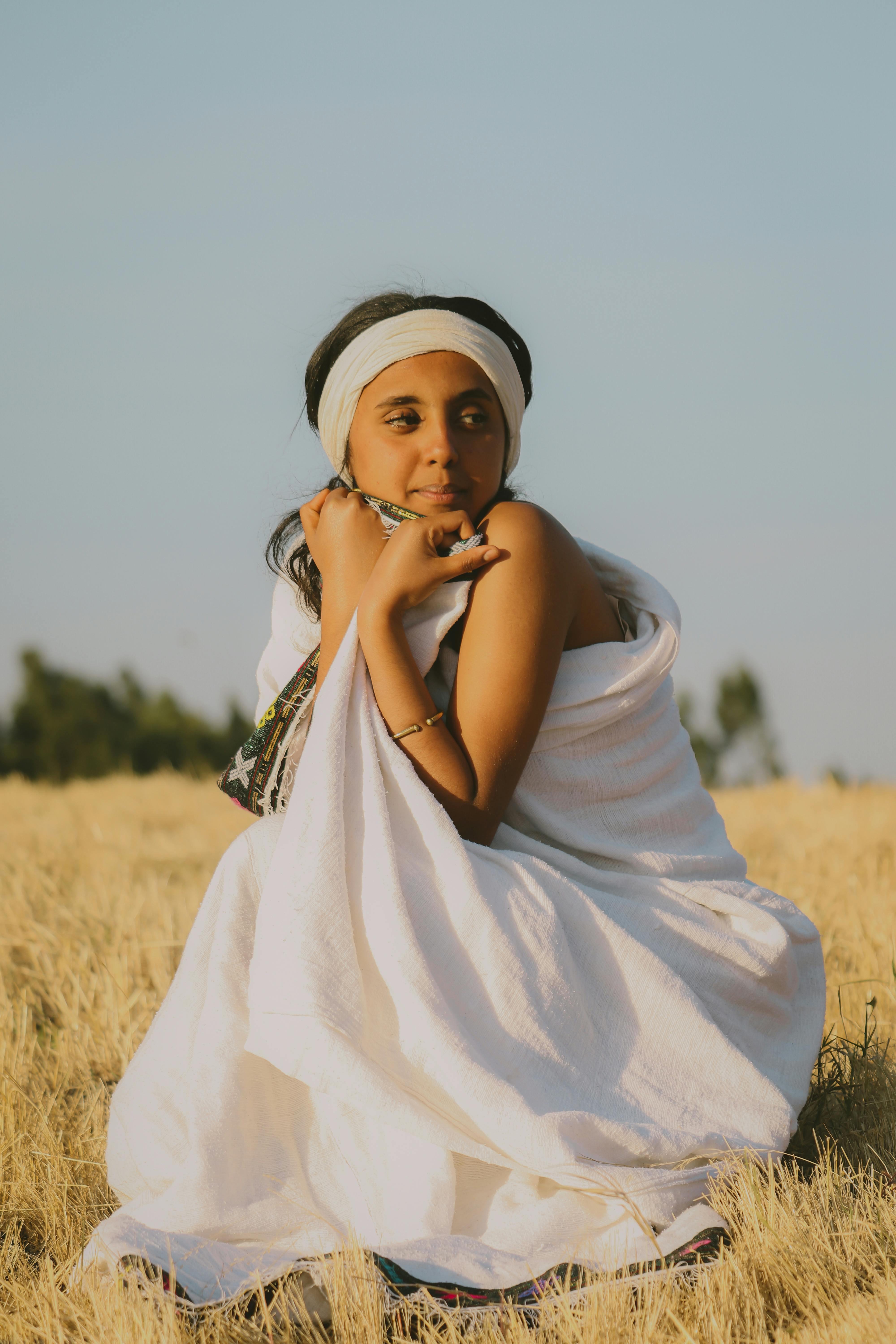 A woman in a white dress poses gracefully in a serene meadow under a clear sky.