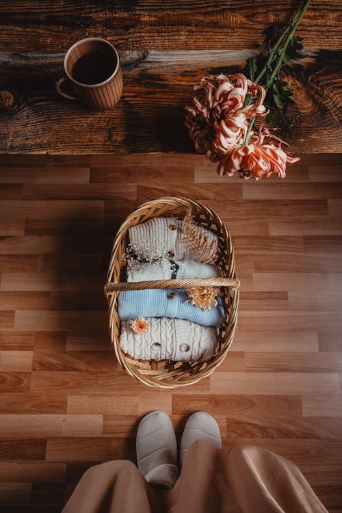 Wicker Basket with Woolen Sweaters Under a Table with a Mug of Coffee and Flowers